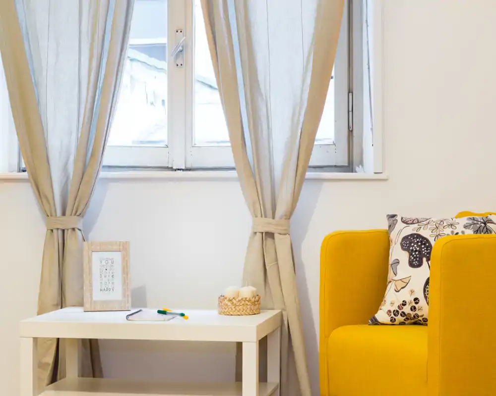 Rincón de sala luminoso y moderno con un sillón amarillo alegre, cojín estampado, cortinas neutras y una pequeña mesa blanca con elementos decorativos — un espacio acogedor junto a la ventana.