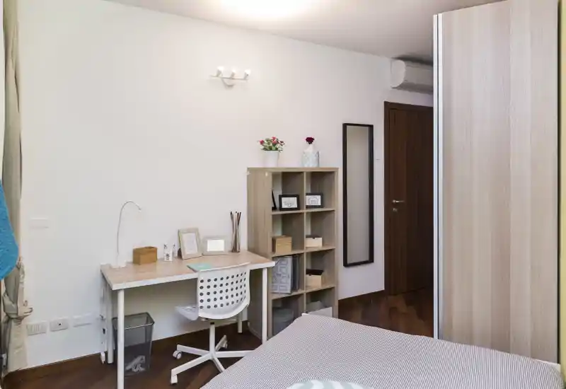 Rincón de dormitorio luminoso y ordenado con cama visible, escritorio y estantería. El ambiente resulta acogedor por los textiles y el almacenamiento organizado, ideal para un anuncio.