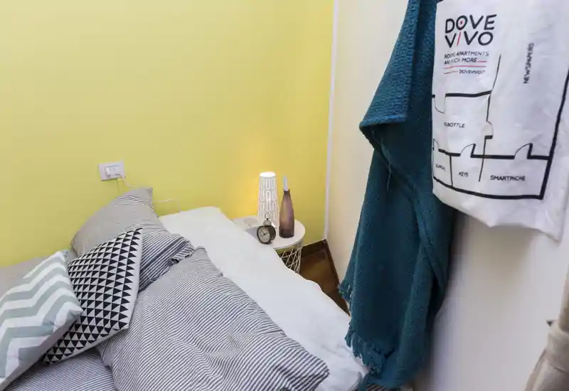 Rincón de dormitorio acogedor y bien decorado con ropa de cama a rayas, cojines estampados y una lámpara de mesita encendida frente a una pared amarilla.