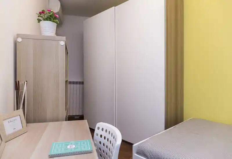 Pequeño rincón de dormitorio ordenado con cama individual, armario y escritorio; luminoso, minimalista y práctico, ideal para estudiantes o estancias cortas.