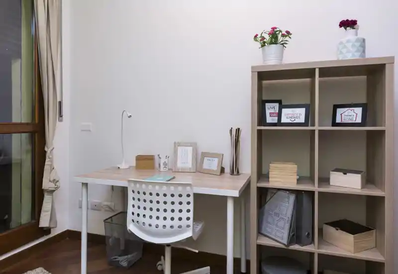 Bright, tidy home workspace with a simple desk, chair and open shelving; decorative frames and plants create a comfortable living-corner feel.