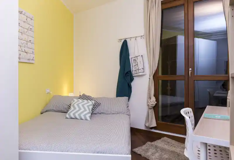 Dormitorio acogedor y bien decorado con cama doble junto a una pared amarilla, cojines decorativos, un pequeño escritorio junto a la ventana y iluminación suave — ideal para una estancia cómoda.