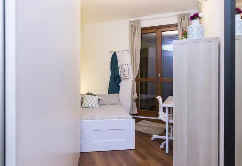 Dormitorio individual acogedor y bien iluminado con cama bien hecha, cojines decorativos y un escritorio compacto junto a la ventana — ideal para estudiante o persona sola.