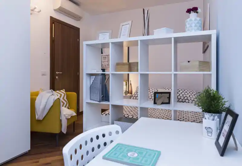 Angolo camera luminoso e ben curato separato da una libreria a giorno bianca; biancheria a motivo, lampada e decorazioni creano un’area notte accogliente e funzionale.