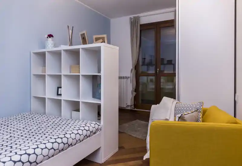 Angolo camera moderno e luminoso con letto singolo ben rifatto separato da un modulo scaffale bianco a cubi e un divano giallo senape che aggiunge un tocco caldo.