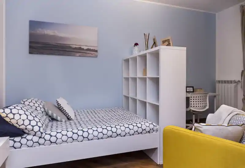 Camera da letto luminosa e ben arredata con letto matrimoniale, biancheria a motivi e una libreria bianca bassa che separa la zona notte dall'angolo lavoro.