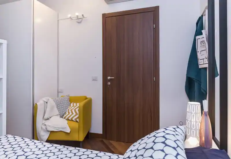 Angolo camera accogliente con biancheria dal motivo visibile in primo piano, una poltrona giallo senape con cuscini e plaid e una porta in legno. Lampada da comodino e allestimento curato creano un'atmosfera luminosa e invitante.