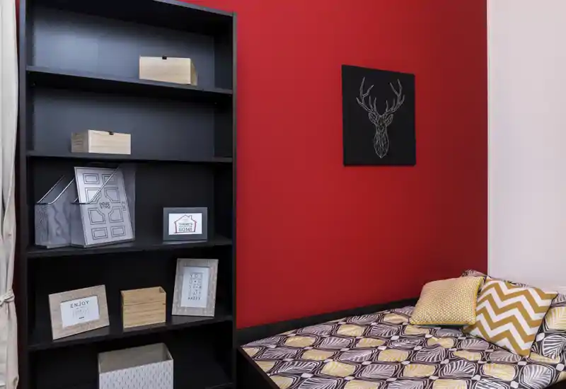 Angolo camera accogliente con biancheria a motivi e cuscini decorativi su una parete rossa accentata, affiancato da una libreria nera alta per riporre oggetti.