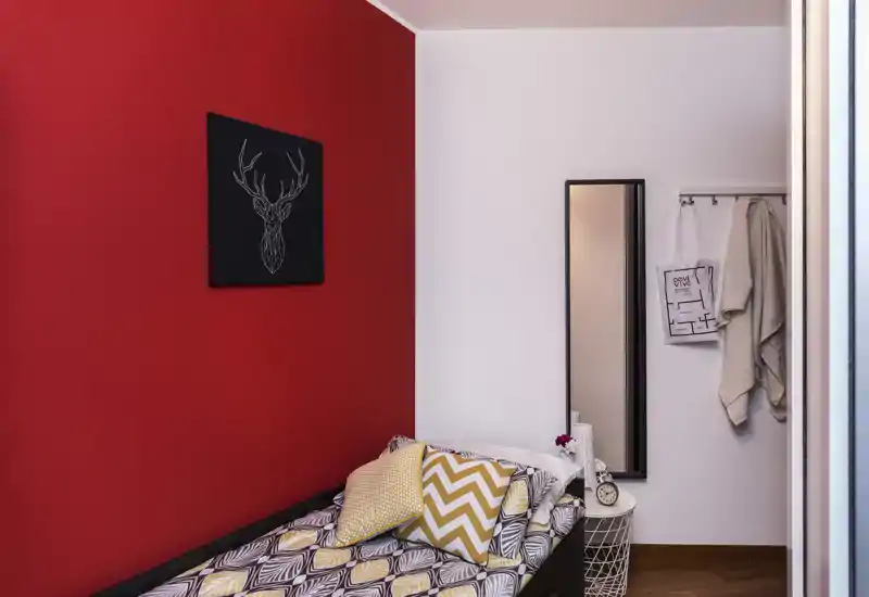 Piccola camera arredata con letto singolo con stile, parete rossa a contrasto, cuscini decorativi e uno specchio a tutta altezza vicino a un appendiabiti.