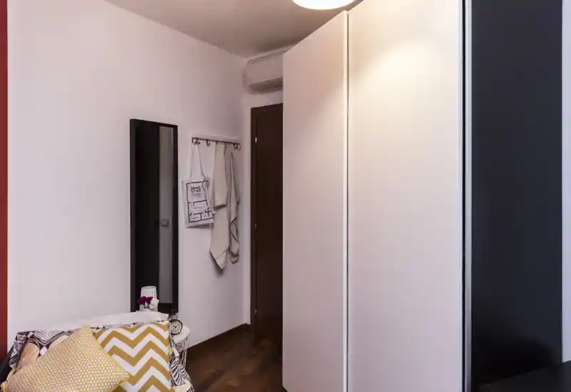 Angolo di camera da letto con letto parzialmente visibile con cuscini decorativi, uno specchio alto, appendini a parete con asciugamani e un armadio bianco — luminoso, ordinato e ben curato.