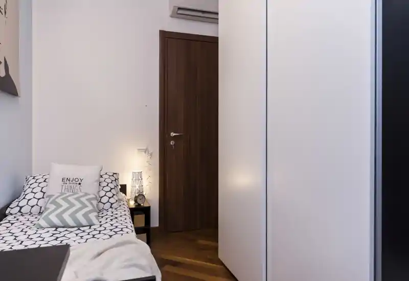 Dormitorio pequeño y acogedor con una cama individual bien hecha, cojines decorativos y una lámpara de mesita. El suelo de madera y la luz suave crean un ambiente cálido y agradable.