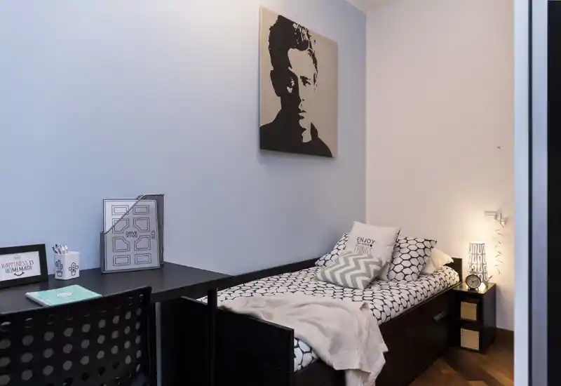 Pequeña habitación decorada con cama individual, ropa de cama estampada y cojines decorativos; escritorio y lámpara crean un rincón acogedor y funcional.