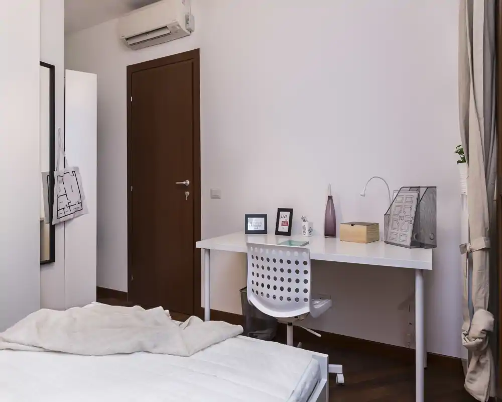 Camera da letto moderna e luminosa con letto visibile in primo piano e una scrivania ordinata contro la parete; arredi neutri e pavimento in legno creano un ambiente tranquillo e funzionale.