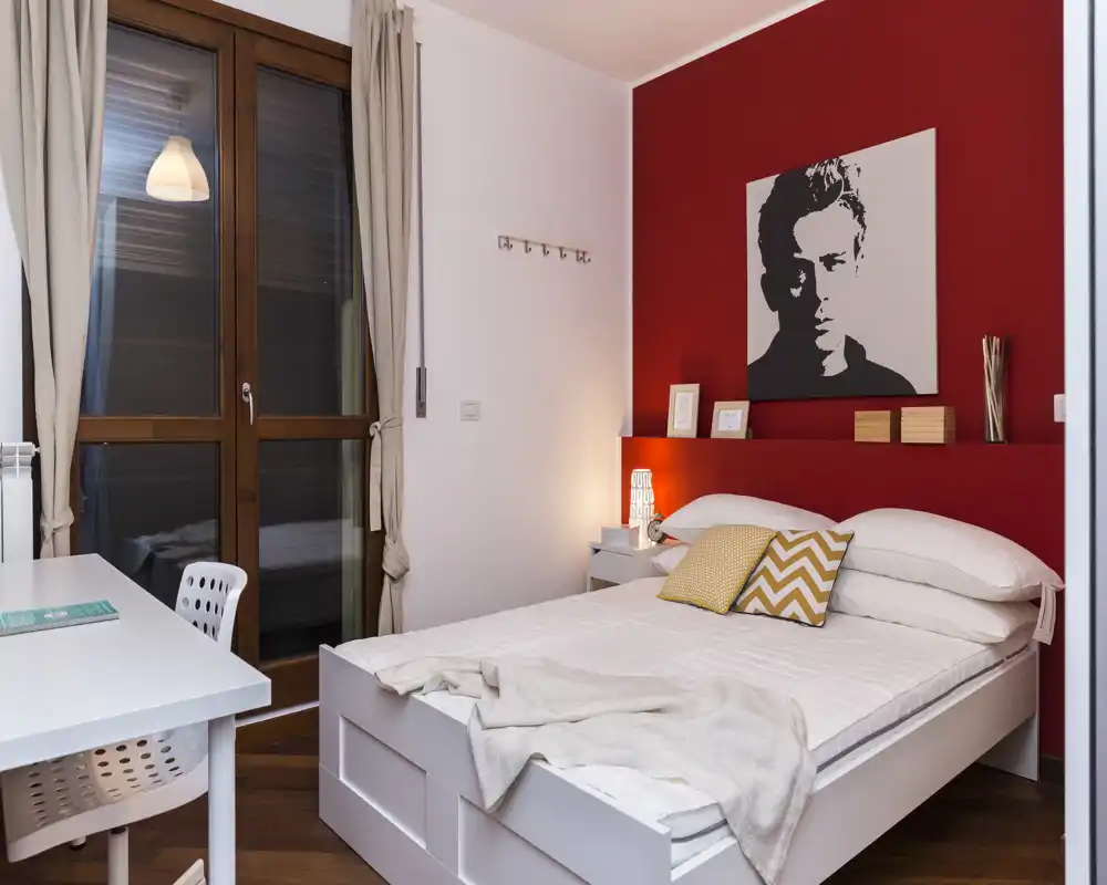 Camera da letto moderna e luminosa con letto bianco, cuscini decorativi e una parete rossa d'accento. Una scrivania vicino alla finestra e l'illuminazione calda del comodino rendono l'ambiente accogliente.