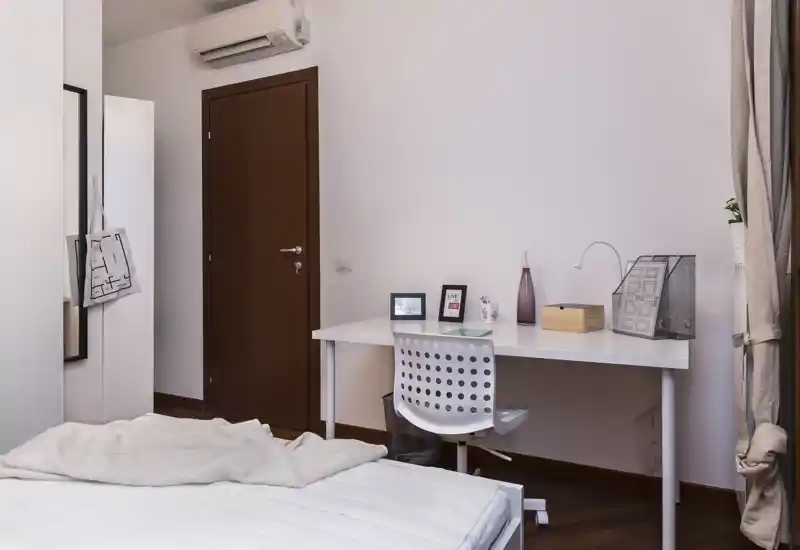 Chambre moderne et lumineuse avec lit visible au premier plan et un bureau rangé contre le mur; décoration neutre et sol en bois pour un espace calme et fonctionnel.