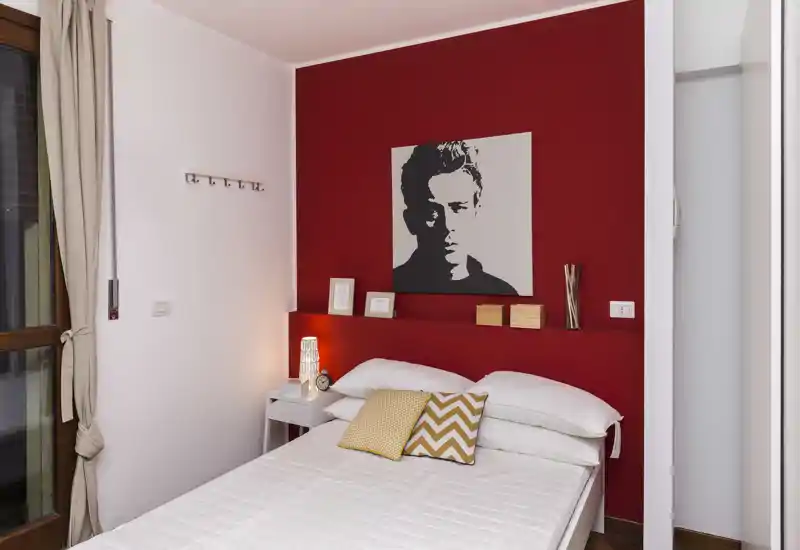 Dormitorio con estilo, cama doble frente a una pared roja intensa, cojines decorativos y una lámpara de noche que crean un ambiente cálido y moderno.