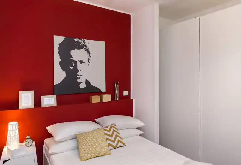 Dormitorio luminoso y con estilo con cama doble frente a una pared roja, cojines decorativos y lámpara de noche; armario moderno a la derecha.