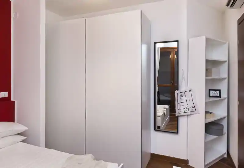 Rincón de dormitorio luminoso y ordenado con cama visible, gran armario blanco, espejo de cuerpo entero y estantería — estilo moderno y minimalista, ideal para anuncios.