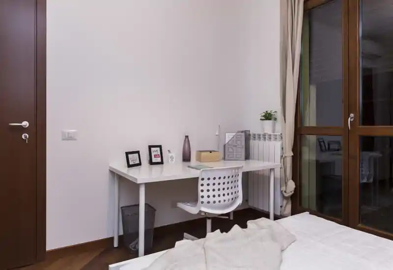 Coin chambre lumineux et soigné avec un bureau blanc, une chaise et un lit bien fait au premier plan ; une grande fenêtre à cadre bois apporte de la lumière naturelle.
