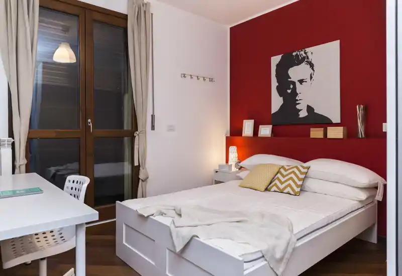 Dormitorio moderno y luminoso con cama blanca, cojines estampados y una llamativa pared roja. Escritorio junto a la ventana y luz de mesita que aportan una atmósfera acogedora.