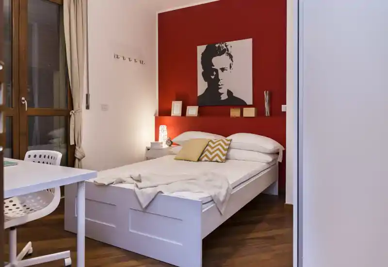 Dormitorio elegante y bien iluminado con cama blanca frente a una pared roja, cojines decorativos y una lámpara de mesita que crean un ambiente acogedor.