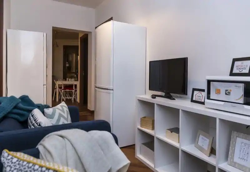 Espace salon chaleureux avec canapé bleu marine, coussins décoratifs et étagère blanche avec TV ; un grand réfrigérateur est visible près de l'entrée vers un coin repas.