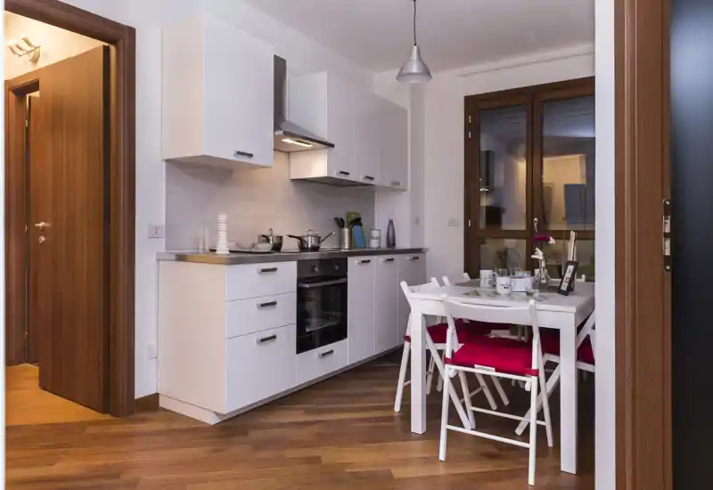Cuisine moderne ouverte avec meubles blancs, four encastré et une petite table à manger équipée de chaises avec coussins rouges — espace lumineux et bien mis en scène, parfait pour un couple ou une petite famille.