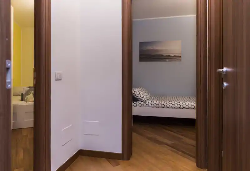 Petite chambre mise en scène visible par l'encadrement de la porte avec un lit simple à linge à motifs, sol en bois et une décoration murale apaisante — espace nuit soigné et prêt à louer.