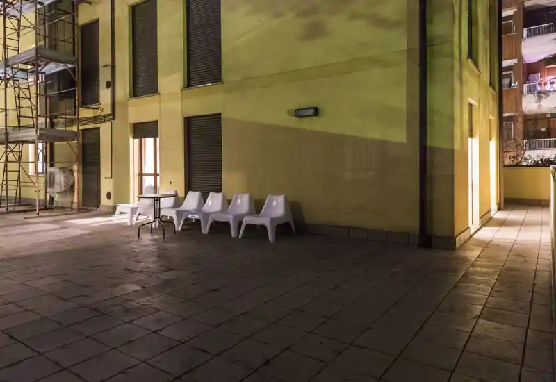 Grande terrasse carrelée la nuit avec des chaises en plastique blanches et une petite table près de la façade jaune de l'immeuble, idéale pour profiter de l'extérieur en soirée.