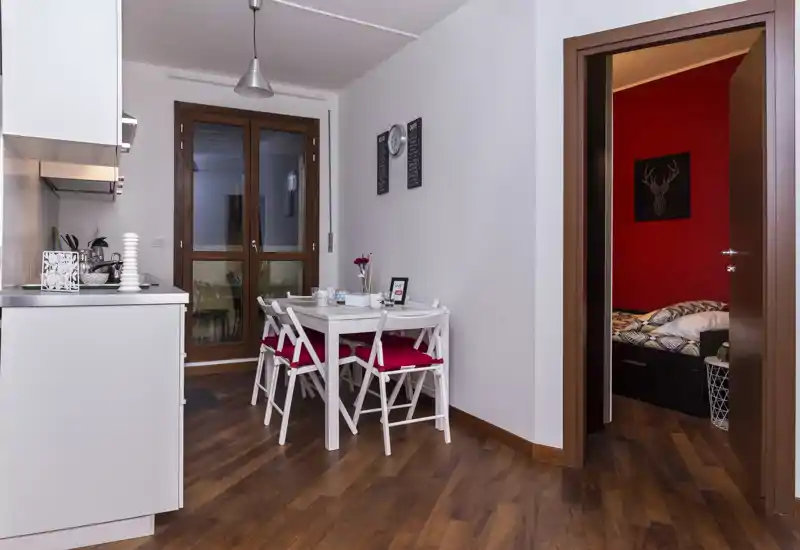 Coin repas compact attenant à la cuisine avec une table blanche et quatre chaises aux coussins rouges, sol en bois et une porte donnant sur une chambre à mur rouge.