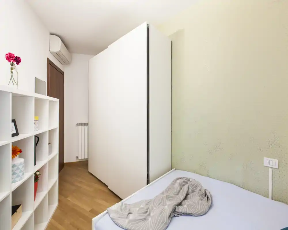 Chambre compacte et moderne avec un lit simple, un mur texturé vert clair et une grande armoire blanche; espace lumineux et bien rangé avec étagères ouvertes et parquet.