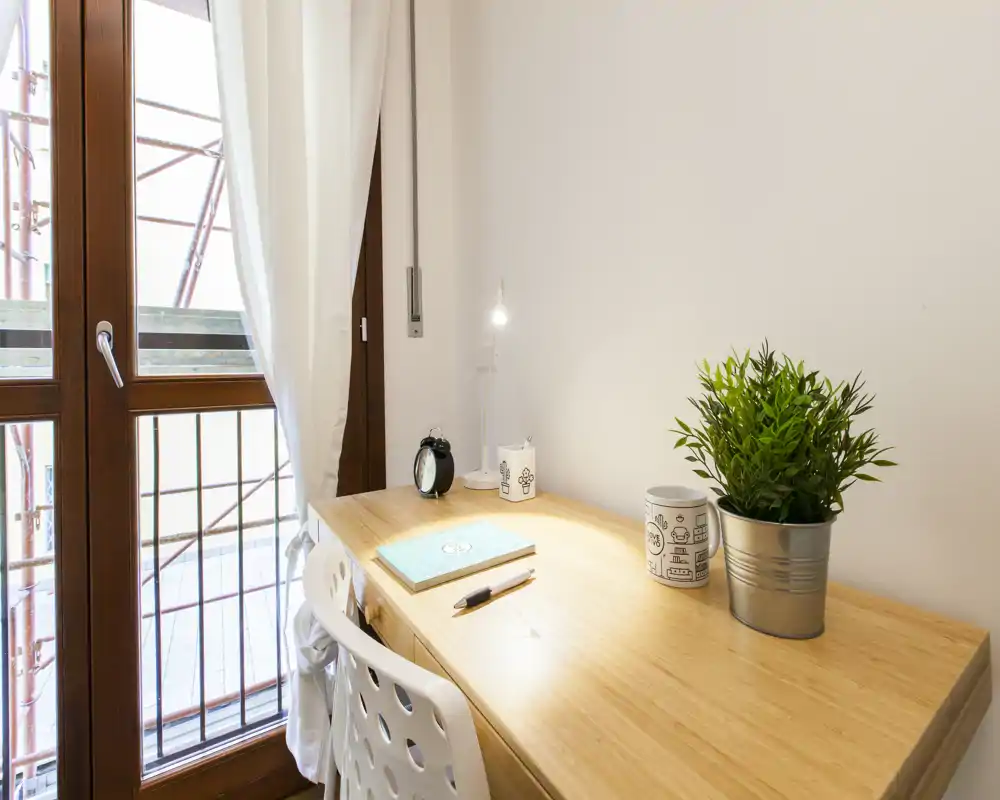 Espace de travail lumineux et soigné près d'une grande fenêtre avec bureau en bois, lampe, petite plante et décoration minimaliste — parfait pour le télétravail ou les études.