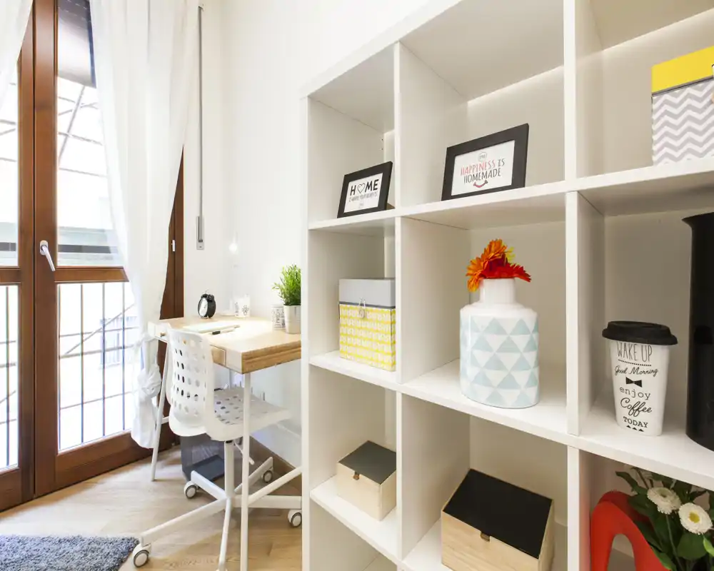 Espace de vie lumineux et soigné avec un coin bureau compact et une étagère modulable. La lumière naturelle de la porte-fenêtre met en valeur le parquet et les accents décoratifs chaleureux.