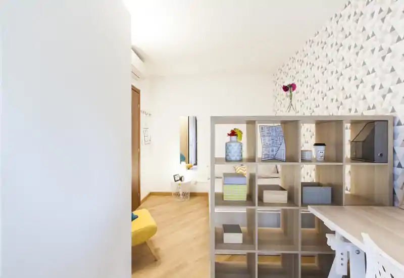 Zona giorno luminosa e moderna con libreria a cubi in legno usata come divisorio, carta da parati decorativa e pavimento in legno caldo — ambiente accogliente e ben arredato perfetto per un annuncio di affitto.