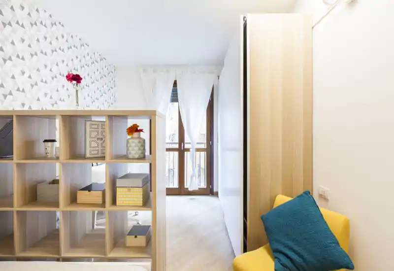 Angolo camera compatto e ben arredato con un divisorio a cubi, una poltrona gialla con cuscino turchese e porta finestra sul balcone che illumina l’ambiente.