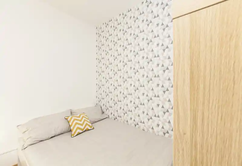 Camera da letto compatta e luminosa con un letto matrimoniale contro una parete decorativa a motivo geometrico e un armadio in legno sul bordo dell’inquadratura. Biancheria neutra e un cuscino giallo a motivo chevron donano un tocco moderno.