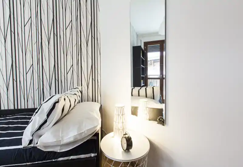 Rincón de dormitorio compacto con cama individual vestida en blanco y negro, mesita con lámpara decorativa y un espejo alto en la pared que refleja la habitación y la puerta del balcón.