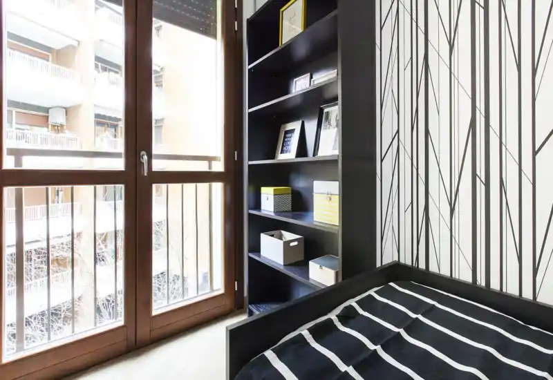 Dormitorio moderno y compacto con cama individual, ropa de cama a rayas, estantería alta y una gran ventana/puerta que aporta abundante luz natural.