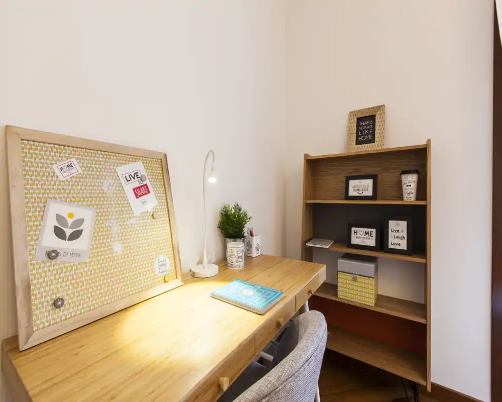 Compatto angolo studio con scrivania in legno, lampada da tavolo, sedia imbottita e libreria con scatole e cornici decorative; luminoso, ordinato e adatto al lavoro da remoto.