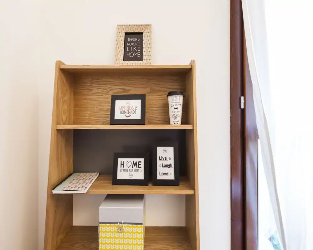 Scaffale decorativo in legno con cornici, una scatola a fantasia e un bicchiere da asporto vicino a una finestra luminosa: un dettaglio d'arredo più che una panoramica della stanza.