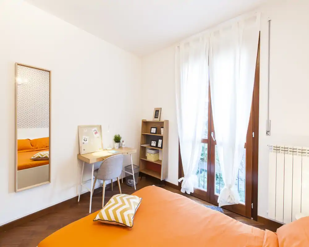 Camera da letto luminosa e moderna con copriletto arancione, cuscino decorativo e una piccola scrivania vicino a una grande finestra che lascia entrare luce naturale.