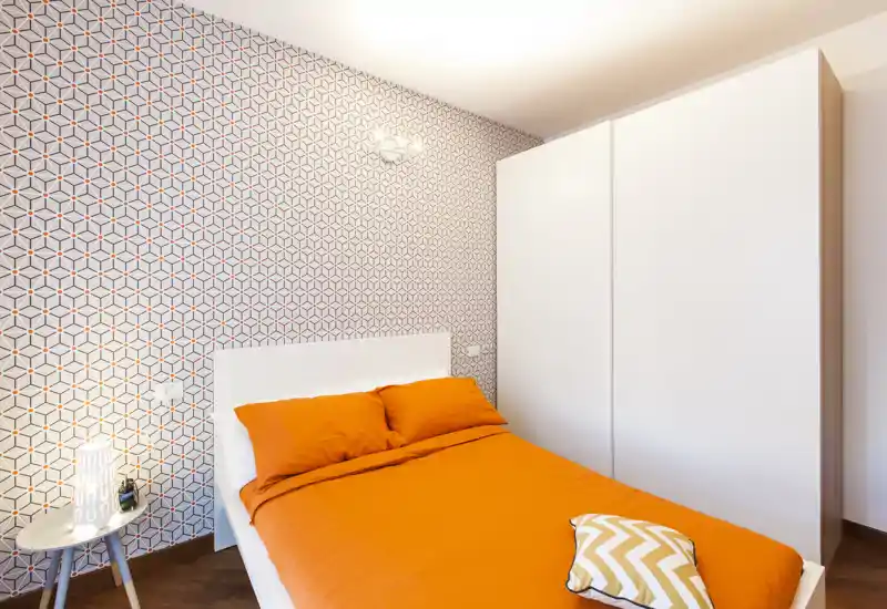 Dormitorio moderno y luminoso con una cama doble cómoda vestida con ropa de cama naranja vivo y una pared geométrica como foco decorativo. Un gran armario blanco y una mesita completan el diseño limpio y contemporáneo.