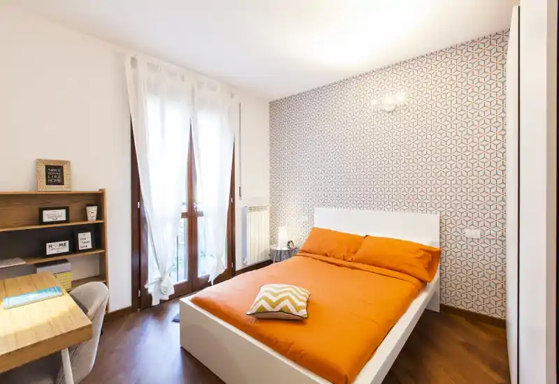 Dormitorio moderno y luminoso con cama doble en ropa de cama naranja, pared de acento con patrón geométrico y un pequeño escritorio junto a la ventana — ideal para alquileres de corta estancia.