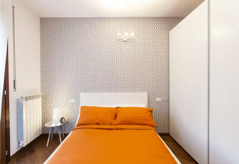 Dormitorio luminoso y ordenado con cama doble con ropa de cama naranja intensa, pared decorativa, mesita con lámpara y armario blanco — moderno y acogedor.
