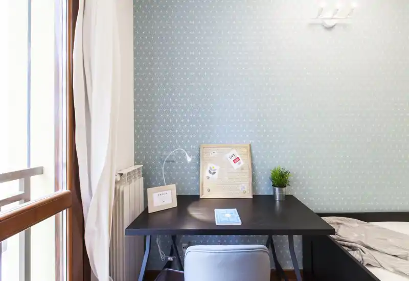 Bright, compact bedroom workspace with a desk next to a window, patterned wallpaper and a partially visible bed. The small desk is styled with a lamp, pinboard and plant — ideal for remote work in a cozy bedroom corner.