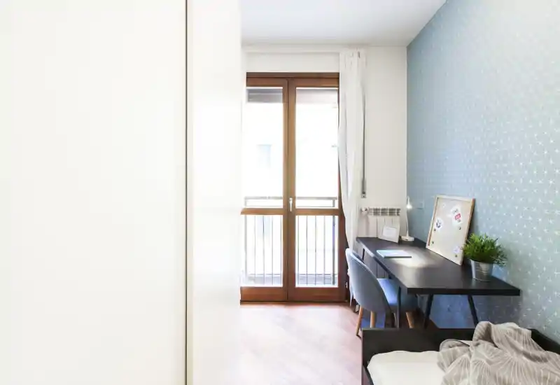 Bright, modern small bedroom with a single bed in the corner, a study desk and chair by a large balcony door; light wood floors and soft patterned wallpaper create a welcoming, functional space.