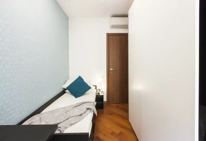 Compact single bedroom with a neatly made bed, teal accent cushion and patterned pale-blue feature wall; wood parquet flooring and warm lighting create a welcoming atmosphere.
