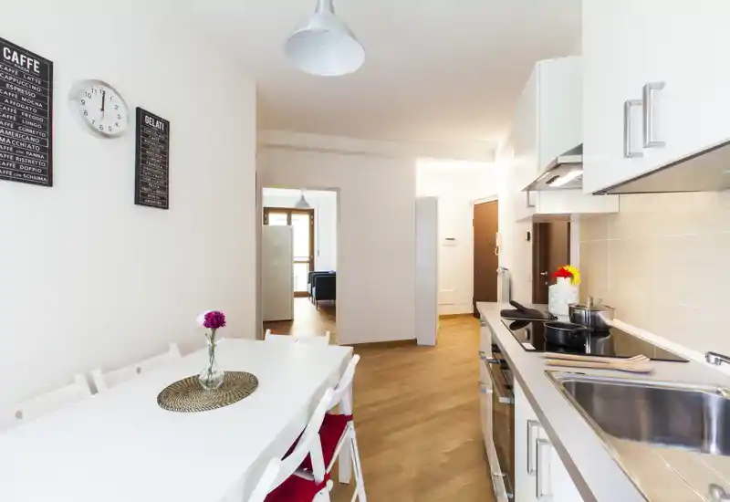 Bright modern kitchenette with integrated appliances and a small dining table; clean white cabinetry and warm wooden floors create a welcoming cooking and dining space.