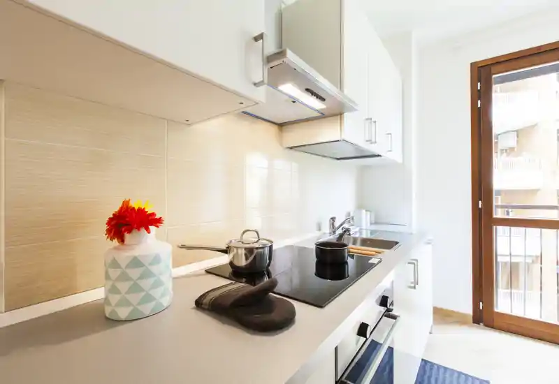 Bright modern kitchen with an induction cooktop, built-in oven and sink along a white countertop; natural light from the balcony door and tasteful decor create a fresh, inviting space.