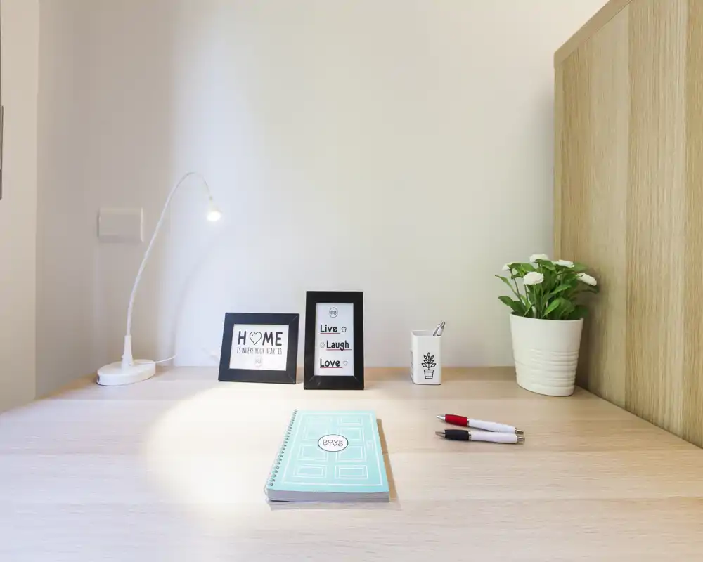 Angolo scrivania ben illuminato in camera da letto con lampada da tavolo, cornici con stampe motivazionali, una piccola pianta e cancelleria — ideale per lavoro o studio.